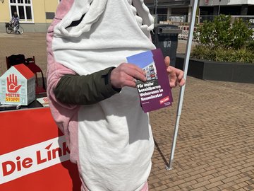 Ein Mensch in einem rosafarbenen Haikostüm verteilt vor einem Pavillon Flyer zum Thema "Bezahlbarer Wohnraum"