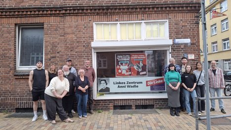 Das Bild zeigt Menschen, die um das Parteibüro der Linken Neumünster herumstehen, in dessen Fenster ein Schild aufgehängt ist. Auf dem Schild steht "Linkes Zentrum Anni Wadle".