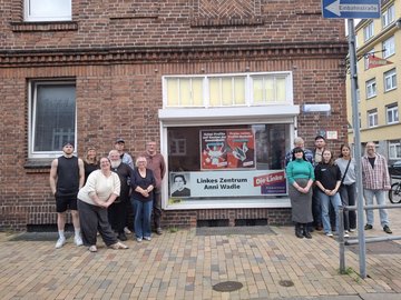 Das Bild zeigt Menschen, die um das Parteibüro der Linken Neumünster herumstehen, in dessen Fenster ein Schild aufgehängt ist. Auf dem Schild steht "Linkes Zentrum Anni Wadle".