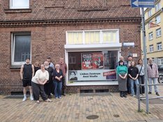 Das Bild zeigt Menschen, die um das Parteibüro der Linken Neumünster herumstehen, in dessen Fenster ein Schild aufgehängt ist. Auf dem Schild steht "Linkes Zentrum Anni Wadle".